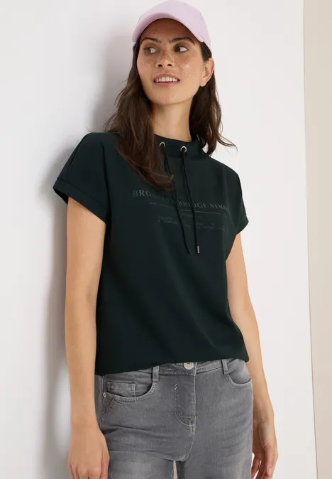 Frau mit dunklem T-Shirt, grauer Jeans und rosa Kappe steht an einer Wand.